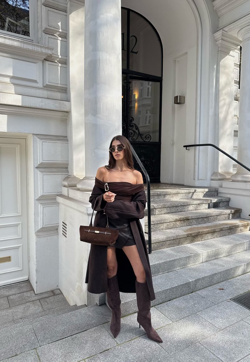 Top marrón oscuro sin hombros, falda de ante a juego y botas hasta la rodilla. Bolso de tela sostenible con acentos dorados, en un entorno urbano elegante.