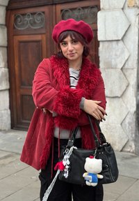 Veste en velours côtelé rouge avec bordure en fausse fourrure, pull rayé gris en dessous, sac à main noir avec porte-clés en peluche, et pantalon foncé.