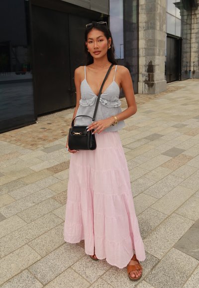 Mujer vestida con una blusa sin mangas a rayas, una falda maxi de volantes rosa, sandalias y sosteniendo un bolso negro en una calle pavimentada urbana.