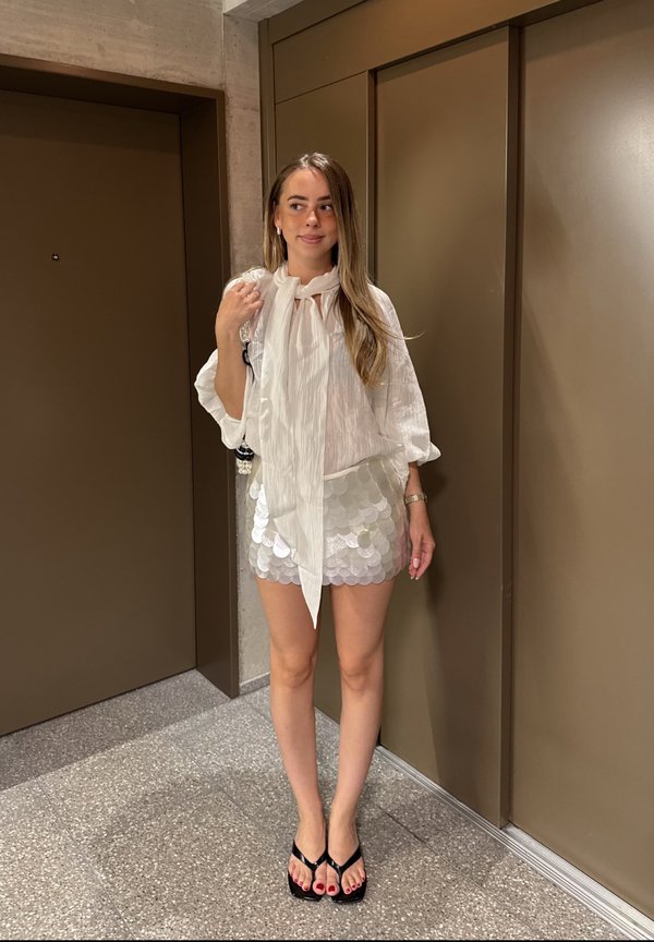 Junge Frau mit langen Haaren steht in einem Flur, trägt eine weiße Bluse, einen silbernen Paillettenrock, schwarze Sandalen und hält eine Handtasche auf der Schulter.