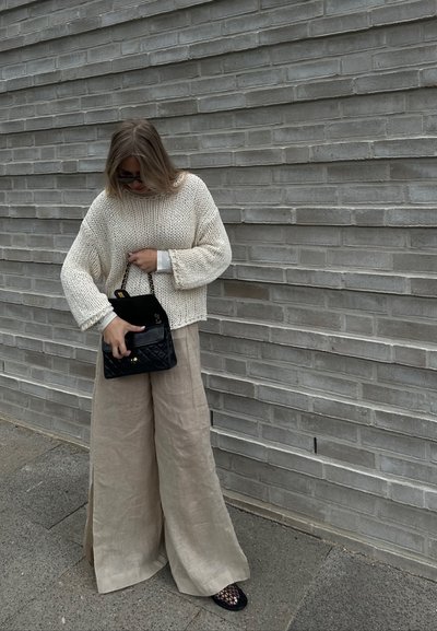 Suéter de punto crema, pantalones de lino anchos sueltos en color beige, bolso negro acolchado y zapatos de malla negros, todo contra una pared texturizada de color gris.