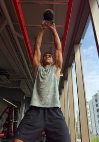 Kettlebell mit Griffen, über dem Kopf gehalten. Nutzer trägt ein graues Tanktop und schwarze Shorts. Fitnessstudio-Umgebung mit großen Fenstern und sichtbarer Trainingsausrüstung.