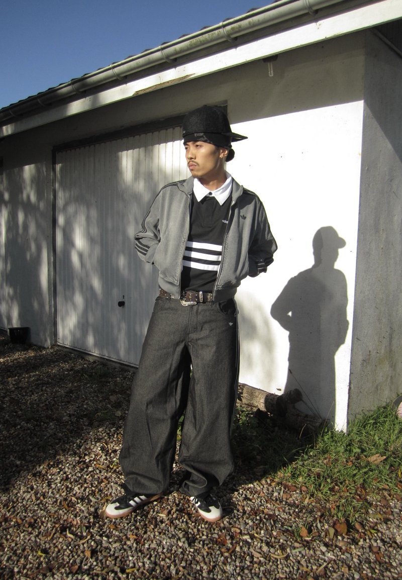 Uomo con cappellino nero, giacca grigia, maglietta bianca e nera, pantaloni larghi scuri e scarpe da ginnastica, in piedi vicino a una porta di garage bianca.