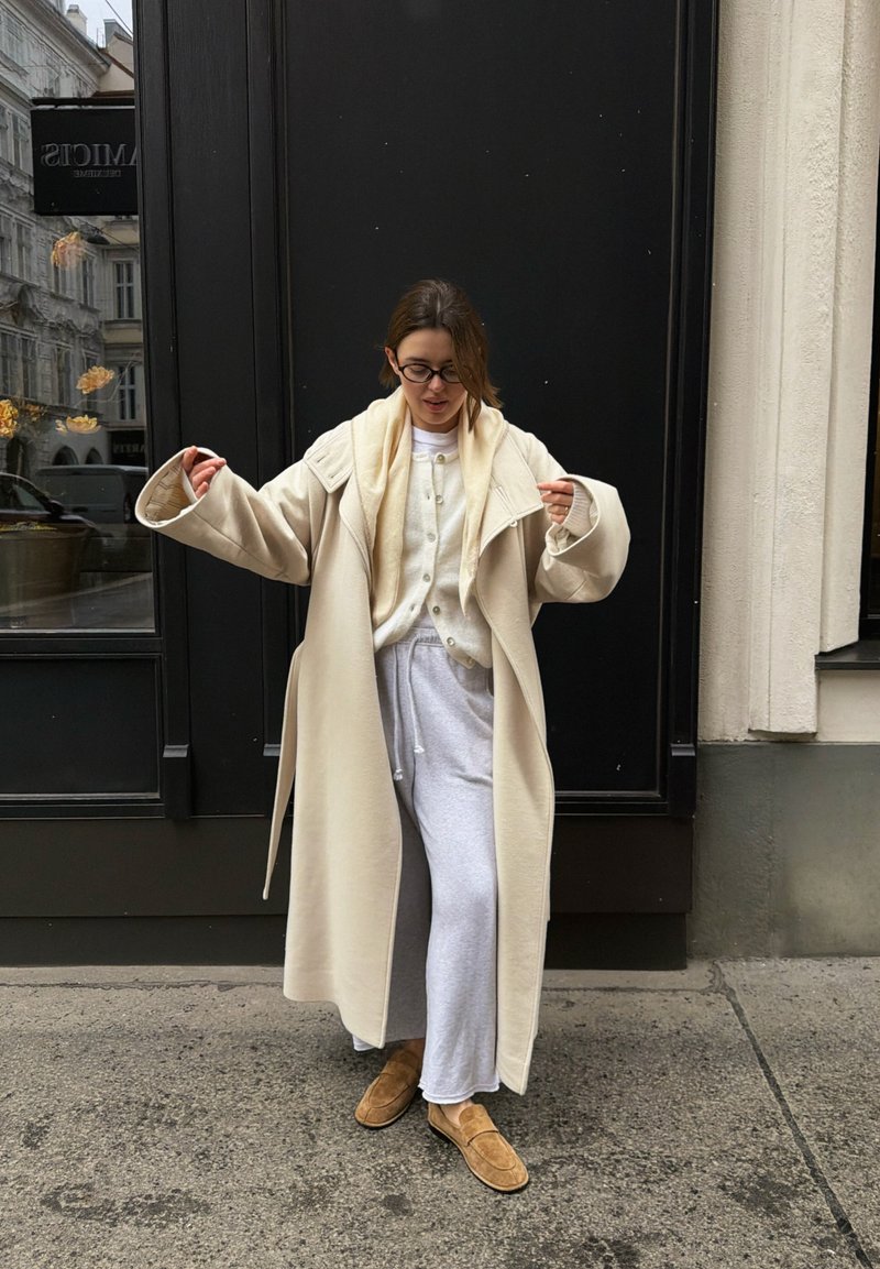 Femme portant un long manteau beige, un cardigan crème, un pantalon de survêtement gris et des mocassins marron, debout sur un trottoir en ville contre une porte noire.