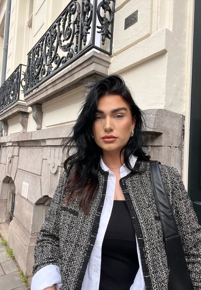 Chaqueta texturizada en blanco y negro con cuello redondo, llevada sobre una blusa negra, combinada con una camisa blanca de cuello, sosteniendo un bolso negro.