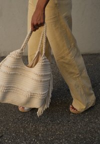 Geflochtener Cremefarbenes Tasche mit strukturierten Streifen und Fransen, getragen von einer Hand. Kombiniert mit hellgelben Hosen und beigen Sandalen.