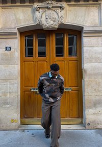 Homme portant un pull camouflage et un pantalon brun marchant devant une porte en bois ornée avec un visage sculpté en pierre au-dessus.
