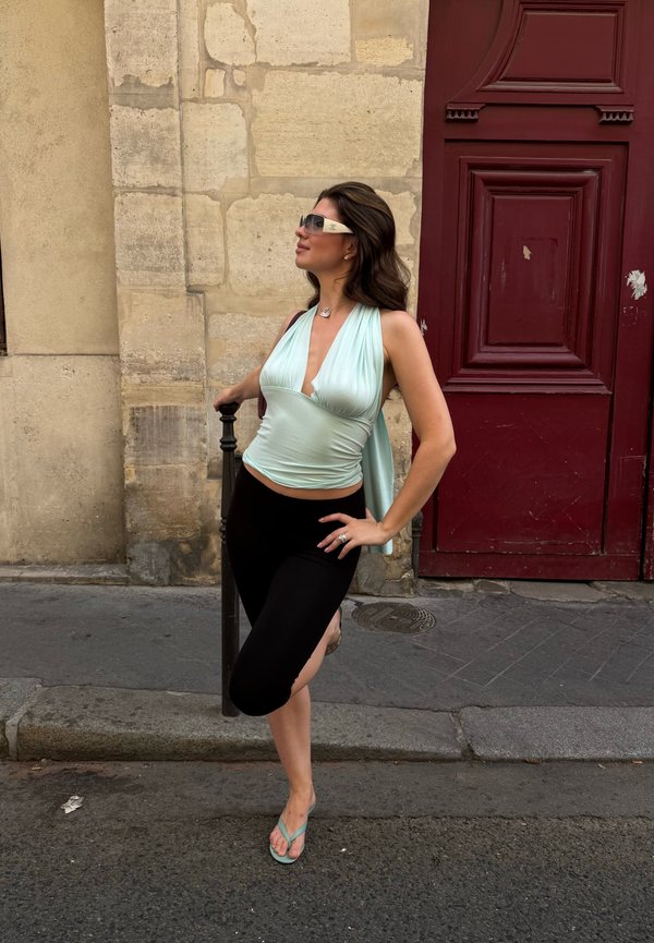 Woman in sunglasses and light blue top leaning on a black post in front of a stone wall and red door on a city street.