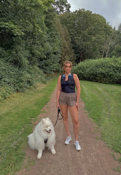 Perro Samoyedo con una correa al lado de una persona que lleva un chaleco negro y pantalones cortos beige. Se pueden ver árboles verdes y un sendero en el fondo.