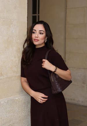 Joven mujer con cabello largo y oscuro con vestido burdeos apoyada contra una pared de piedra en el interior, llevando un bolso de cuero tejido en el hombro.