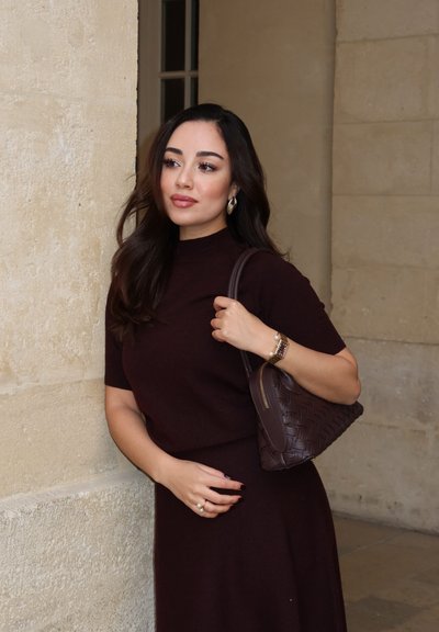 Joven mujer con cabello largo y oscuro con vestido burdeos apoyada contra una pared de piedra en el interior, llevando un bolso de cuero tejido en el hombro.