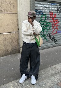 Bílá bomber bunda, tmavé oversized džíny a bílé tenisky. Drží zeleno-béžovou tašku, stojí proti zdi pokryté graffiti.