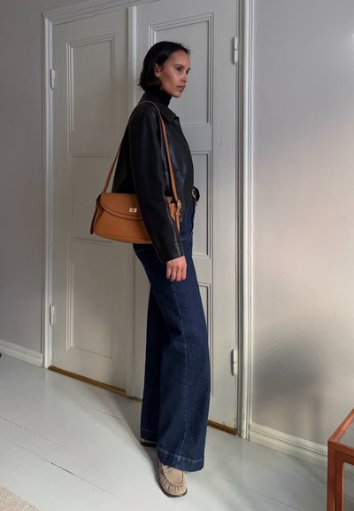 Mujer de pie en interiores, con chaqueta de cuero negra, jeans anchos azul oscuro, zapatos beige y un bolso de mano marrón claro colgado del hombro.