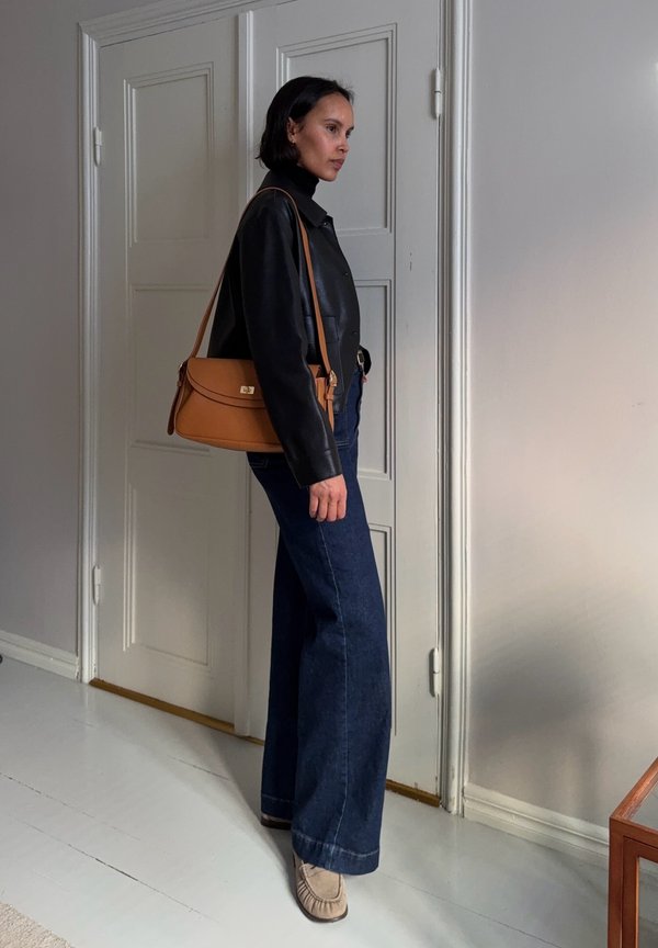 Frau steht drinnen, trägt eine schwarze Lederjacke, dunkelblaue Jeans mit weitem Bein, beige Schuhe und hat eine braune Handtasche über der Schulter.