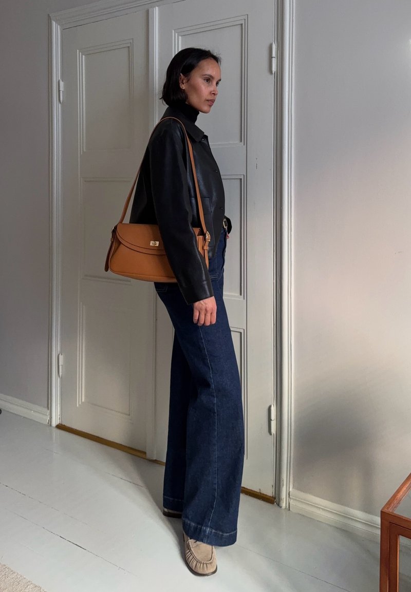 Sieviete, kas stāv telpās, tērpusies melnā ādas jakā, tumši zilos platu kāju džinsos, ar bēšām kurpēm un pār plecu nēsā tumši brūnu somiņu.