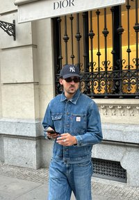 Džínová bunda s knoflíkovým zapínáním, dvěma předními kapsami a modrým odstínem. V kombinaci s odpovídajícími džíny, černou čepicí a tmavými slunečními brýlemi. Moderní městské oblečení.