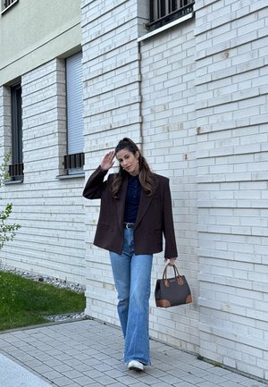 Mujer caminando por un camino pavimentado junto a un edificio de ladrillo blanco, vistiendo una chaqueta oscura, vaqueros azules, zapatos blancos y llevando un bolso.