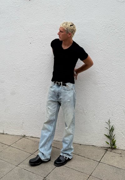 Camiseta negra ajustada, jeans claros desgastados con parches y zuecos negros. Textura suave y ajuste casual contra una pared blanca texturizada.