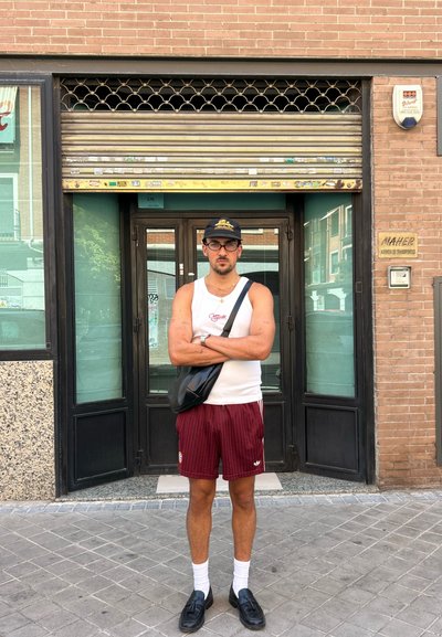 Hombre con una camiseta sin mangas blanca y pantalones cortos a rayas burdeos se encuentra frente a una tienda con una persiana metálica. Sostiene una bolsa negra.