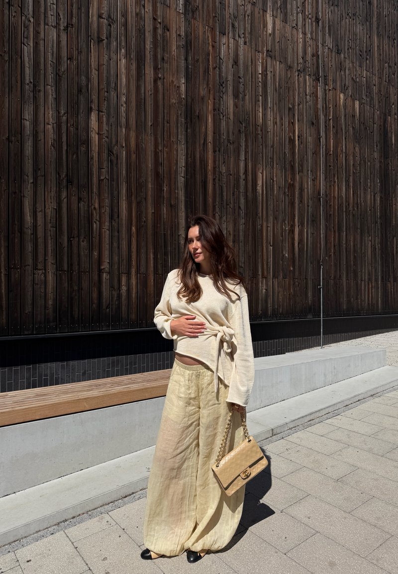 Femme debout sur le trottoir portant un pantalon large beige et un pull, tenant un sac à main beige, avec un grand mur en panneaux de bois foncé derrière elle.