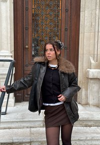 Chaqueta de cuero marrón con cuello de piel, superpuesta a un suéter negro abotonado y una camisa blanca, combinada con una falda marrón y medias.