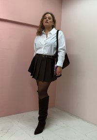 Biała koszula zapinana na guziki włożona w plisowaną brązową spódnicę, zestawiona z brązowymi botkami do kolan. Dodatki obejmują brązową torbę i breloczek.