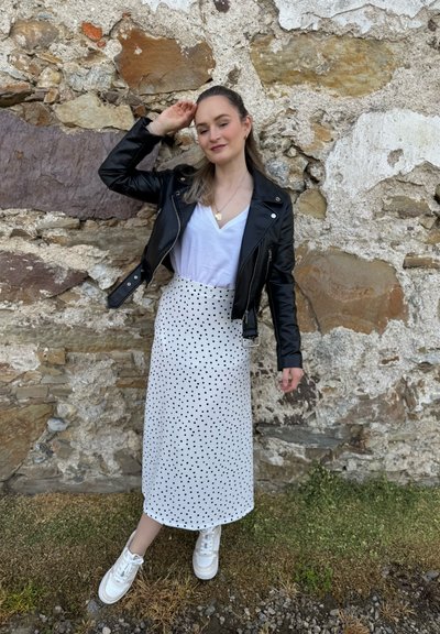 Chaqueta de motorista de cuero negro sobre una camiseta blanca de escote en V, combinada con una falda midi blanca con lunares y zapatillas blancas contra una pared de piedra.