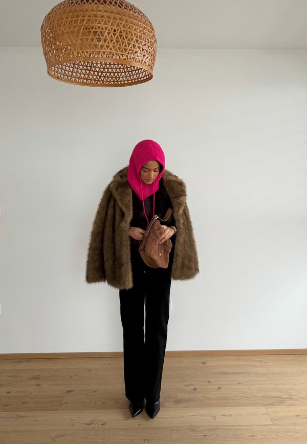 Donna che indossa un cappotto di pelliccia marrone, passamontagna rosa lavorato a maglia, outfit nero, che tiene una borsa marrone, in piedi su un pavimento in legno chiaro sotto una lampada a soffitto intrecciata.