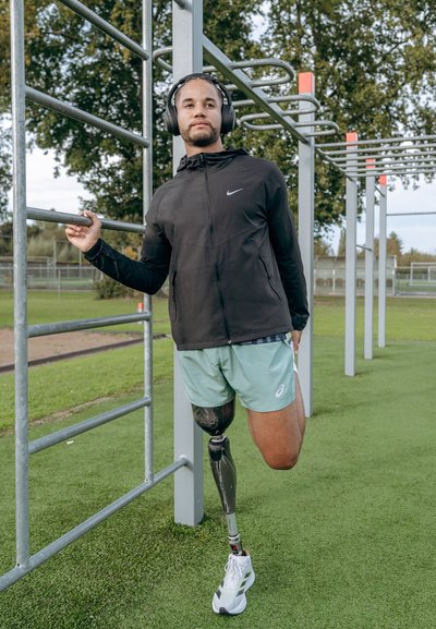 Atleta con chaqueta negra de Nike y pantalones cortos verde claro, usando una barra horizontal de metal, presenta una pierna prostética y auriculares inalámbricos.