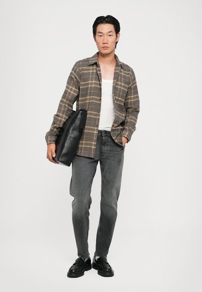 Camisa de cuadros marrón y beige, camiseta sin mangas blanca, jeans grises, sosteniendo un bolso de cuero negro. Zapatos negros con suelas gruesas. Fondo neutro.