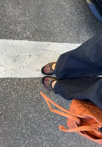 Ballerines en mesh noir avec orteils ouverts, pantalon large en denim noir, et un sac tissé orange sur une surface en asphalte gris.