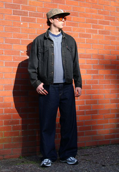 Chaqueta de mezclilla negra sobre una camiseta de rayas azul y blanca, combinada con vaqueros azules holgados y zapatillas grises. Los accesorios incluyen una gorra estampada y gafas de sol naranjas.