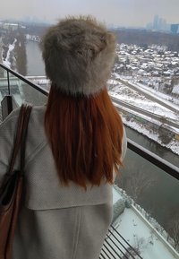 Personne aux longs cheveux roux portant un chapeau en fourrure se tient sur un balcon surplombant une rivière enneigée et un quartier résidentiel en hiver.