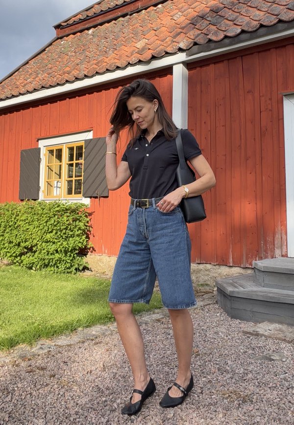 Donna con camicia nera e pantaloncini di jeans che sta su un sentiero di ghiaia vicino a una casa di legno rossa con tetto di tegole e un cespuglio verde.