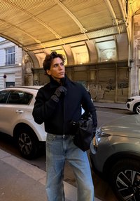 Melna jaka ar rāvējslēdzēju, gaiši zilas taisnas bikses, melnas cimdi un melna soma. Ielas vide ar automašīnām un grafiti fonu.