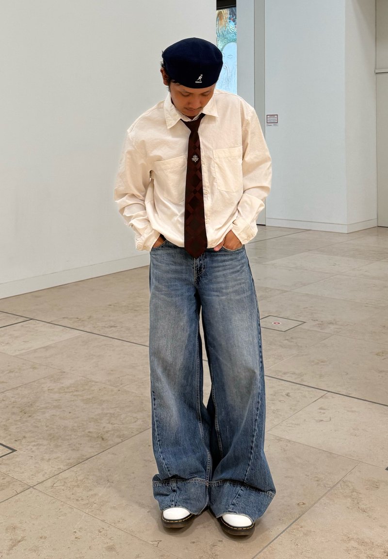 Camicia bianca a maniche lunghe, cravatta a motivo scuro, pantaloni in denim blu oversize, sneakers bianche e berretto blu navy. In piedi all'interno con le mani nelle tasche.