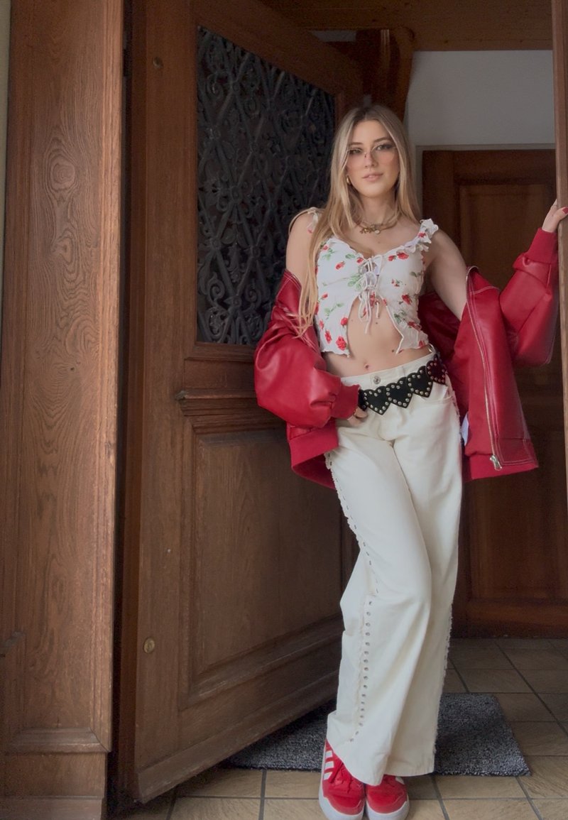Junge Frau lehnt sich an einen hölzernen Türrahmen, trägt ein blumiges Crop-Top, eine rote Jacke von den Schultern gerutscht, weiße Hosen, schwarzen Gürtel und rote Turnschuhe.