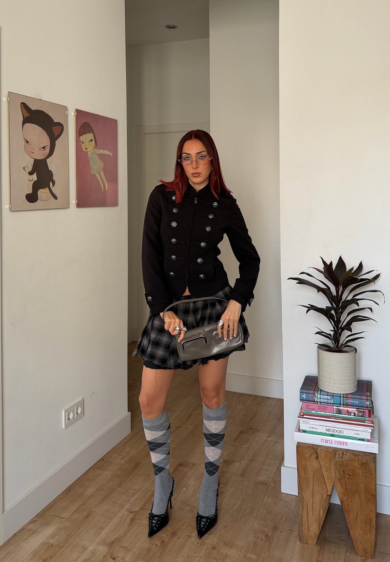 Woman with red hair wears black military-style jacket, plaid skirt, argyle knee socks, and heels, holding a gray clutch bag indoors.