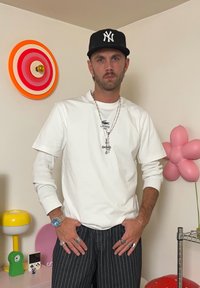Witte lange mouwen t-shirt met een blauw logo, grijs gestreepte broek, zilveren ketting met kruis, zwarte pet en verschillende ringen. Op de achtergrond kleurrijke decoratie.