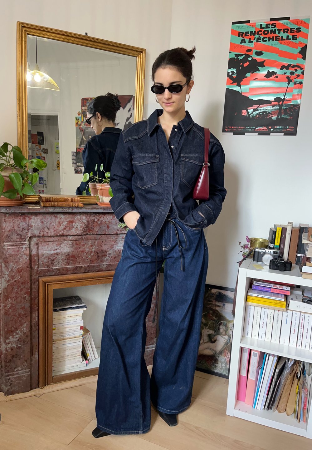 Femme portant une veste en denim foncé et un jean à jambes larges, les mains dans les poches, portant des lunettes de soleil et portant un petit sac rouge, dans une pièce avec des livres et des plantes.