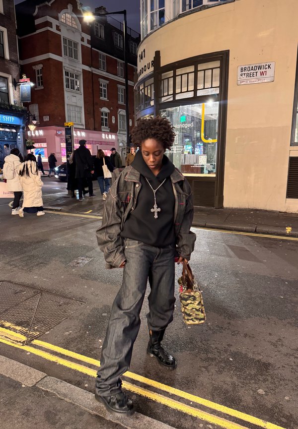 Jeune personne portant une veste et un pantalon en jean foncé, un sweat à capuche noir avec un collier croix, portant un sac camouflage dans une rue de la ville la nuit.