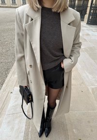 Bézs dupla soros kabát sötét pulóver fölött, fekete rövidnadrággal és térdig érő, fényes fekete csizmával, fekete táskával, arany színű díszítéssel.