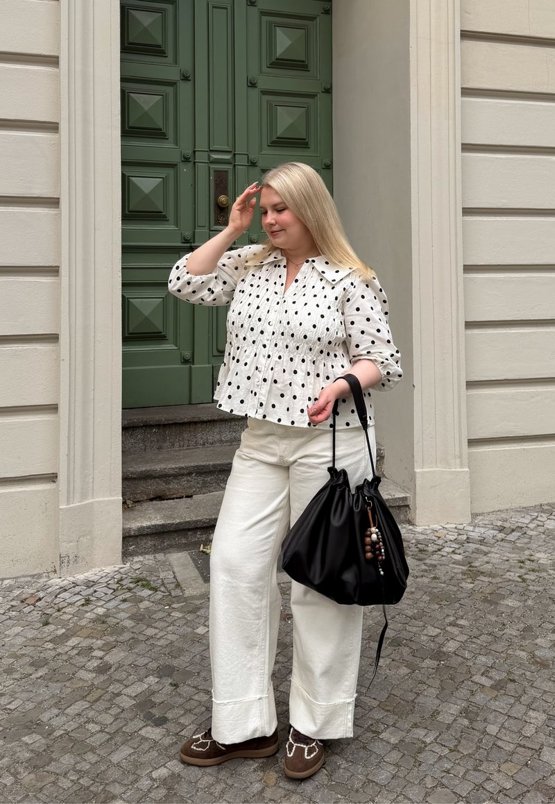 Femme aux longs cheveux blonds portant un chemisier blanc à pois et un pantalon blanc à jambes larges se tient dans une rue pavée en tenant un sac à main noir près d'une porte verte.