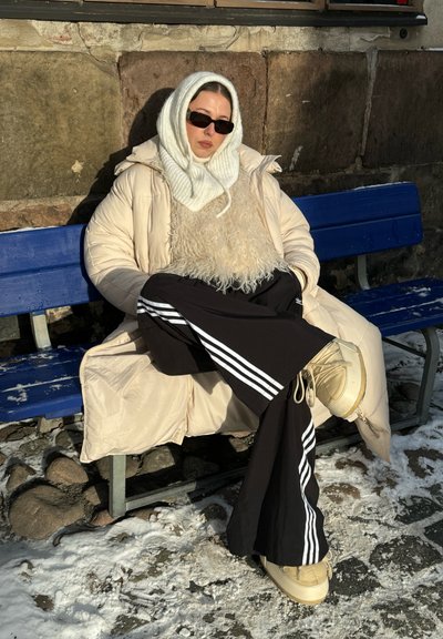 Abrigo de plumas en beige claro con una bufanda de punto blanca y esponjosa, pantalones de chándal negros con rayas blancas y botas de plataforma beige contra un fondo nevado.