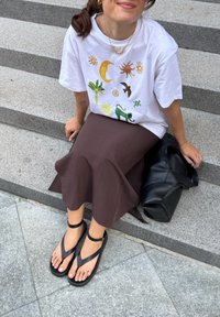 T-shirt branca com designs coloridos de sol, lua e natureza, combinada com uma saia midi castanha e sandálias planas pretas. Bolsa preta ao lado.