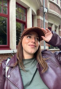 Brun struktureret hat, lilla læderjakke og olivengrøn skjorte, sat imod en murstensbygning med store røde vinduer.