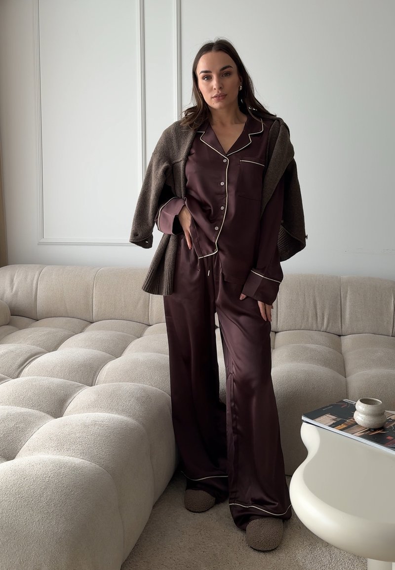 Femme portant un pyjama en satin foncé et un pull marron drapé sur les épaules, debout à côté d'un canapé beige capitonné et d'une table blanche.