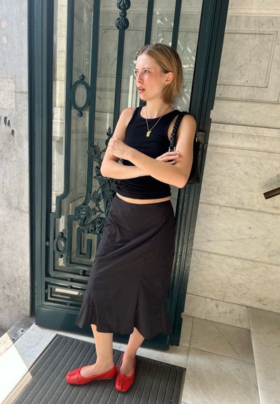 Top sin mangas negro, falda negra con dobladillo acampanado y zapatos planos rojos. Los accesorios incluyen un bolso negro trenzado y un collar con colgante dorado.