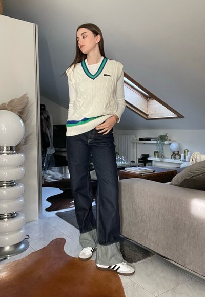 Chaleco de punto blanco con acentos en azul y verde, combinado con jeans de mezclilla oscuros de corte acampanado y zapatillas blancas con rayas negras. Ambiente interior moderno.