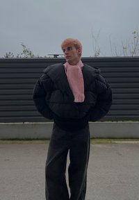 Veste matelassée noire à coupe oversize, portée sur un pull noir, associée à une écharpe rose clair et un pantalon large foncé.